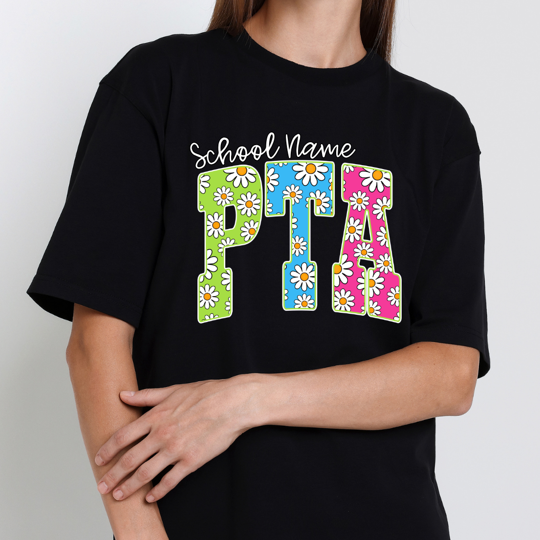 Black t-shirt featuring colorful daisy-pattern PTA lettering in green, blue, and pink with optional school name personalization.