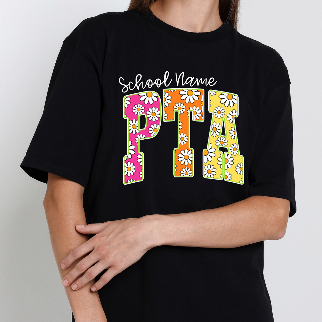 Black t-shirt featuring colorful daisy-pattern PTA lettering in pink, orange, and yellow with optional school name personalization.